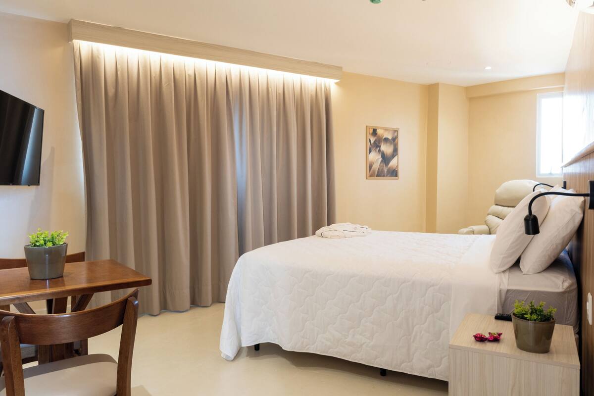 A hotel room with a large bed, a bedside table with a lamp, a TV mounted on the wall, and a window with curtains.
