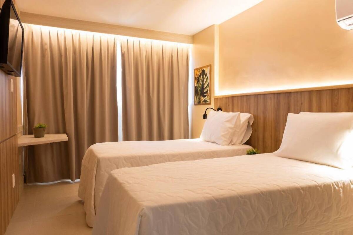 A hotel room with two beds, a TV on the wall, a plant on a shelf, and a large window with curtains.