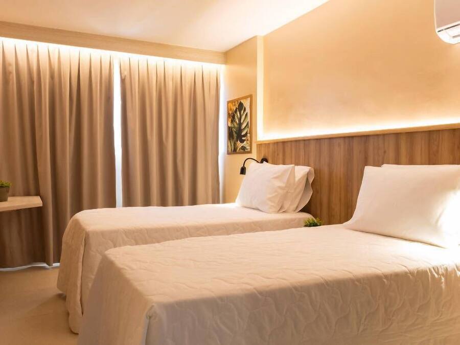 Habitación de hotel con dos camas, un televisor en la pared, una planta en un estante y un ventanal con cortinas.