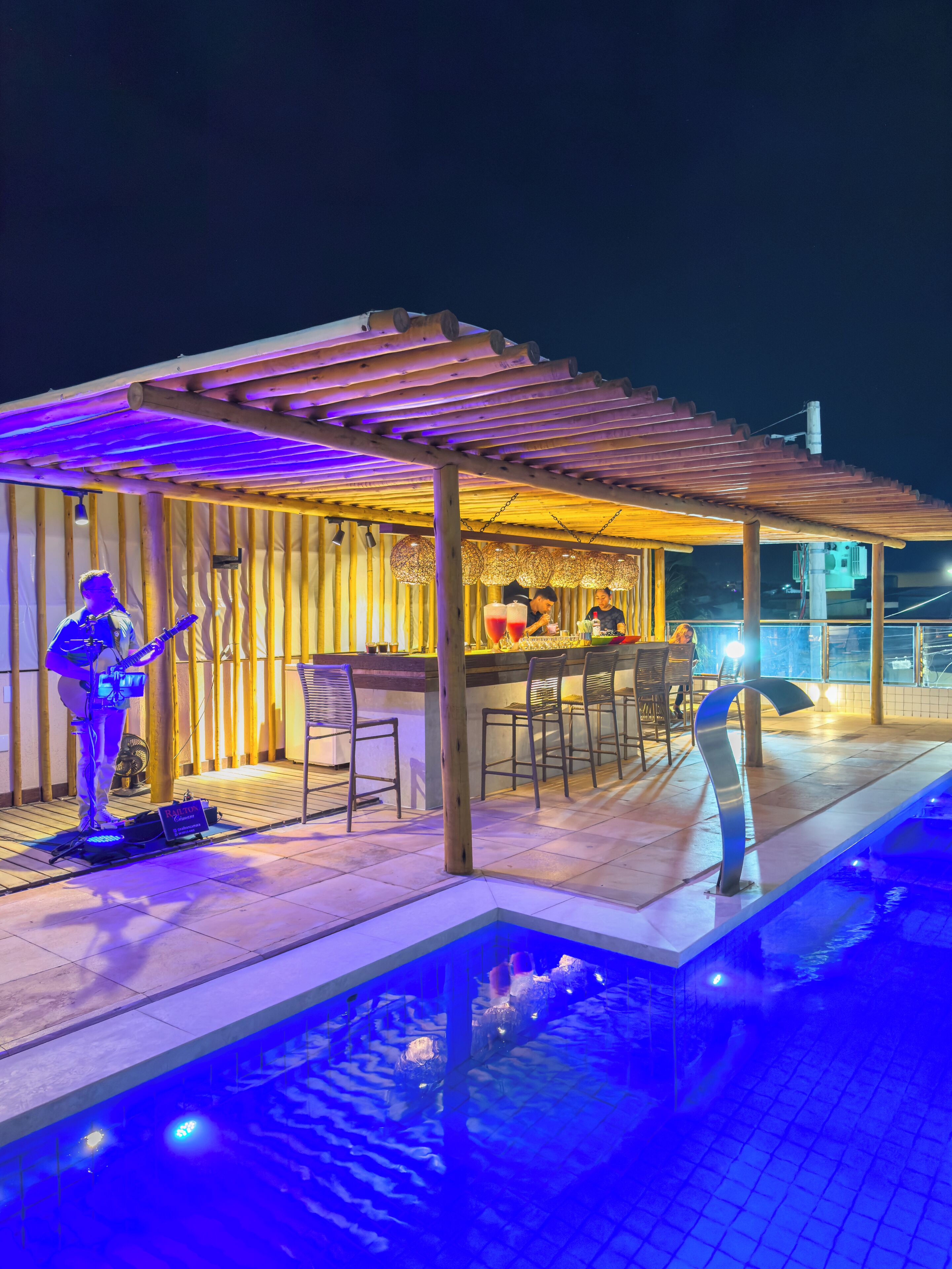 Uma área à beira da piscina com um espaço de estar coberto, um músico e um bar.