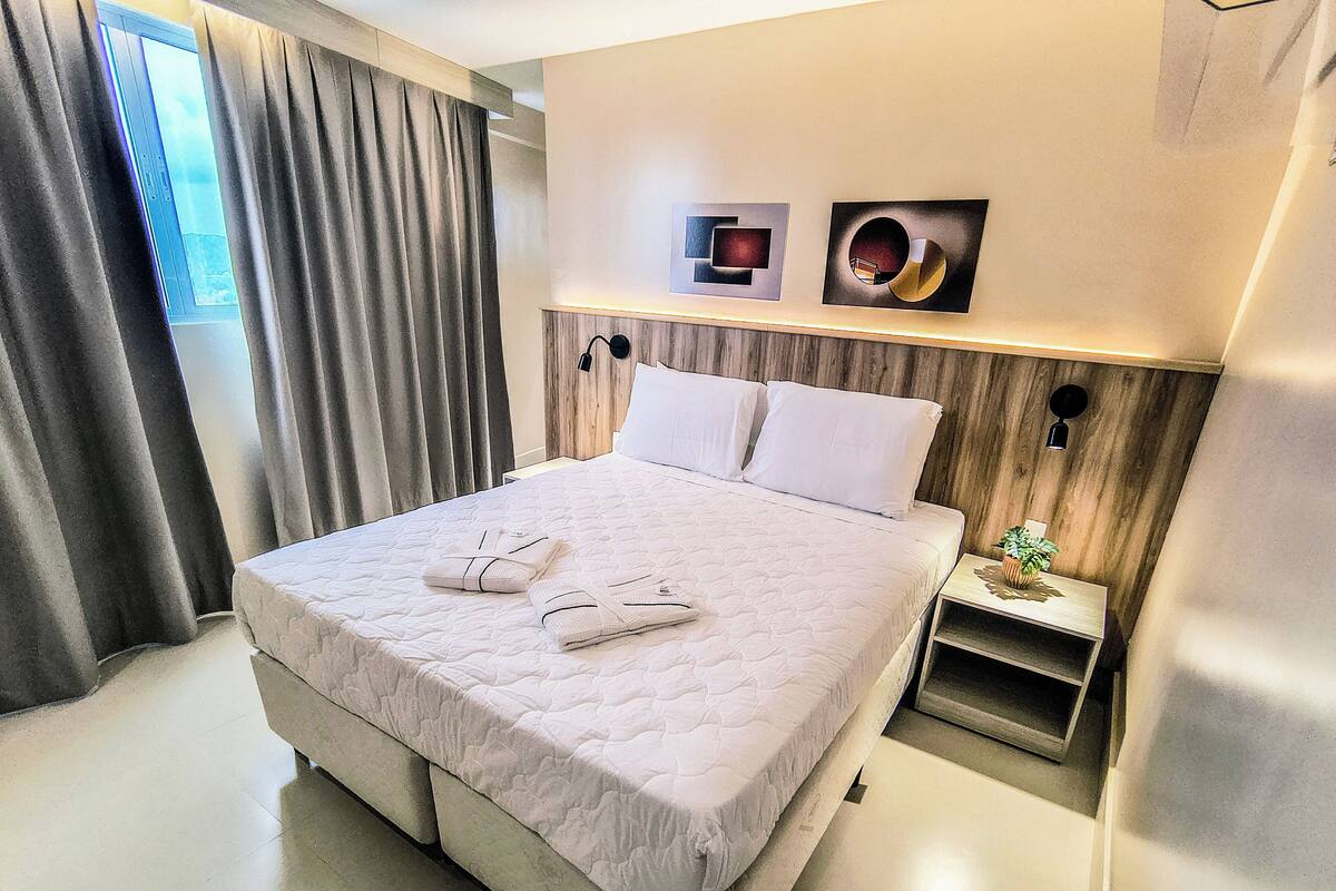 A neatly made bed with pillows and folded towels, a bedside table with a plant, and artwork on the wall.
