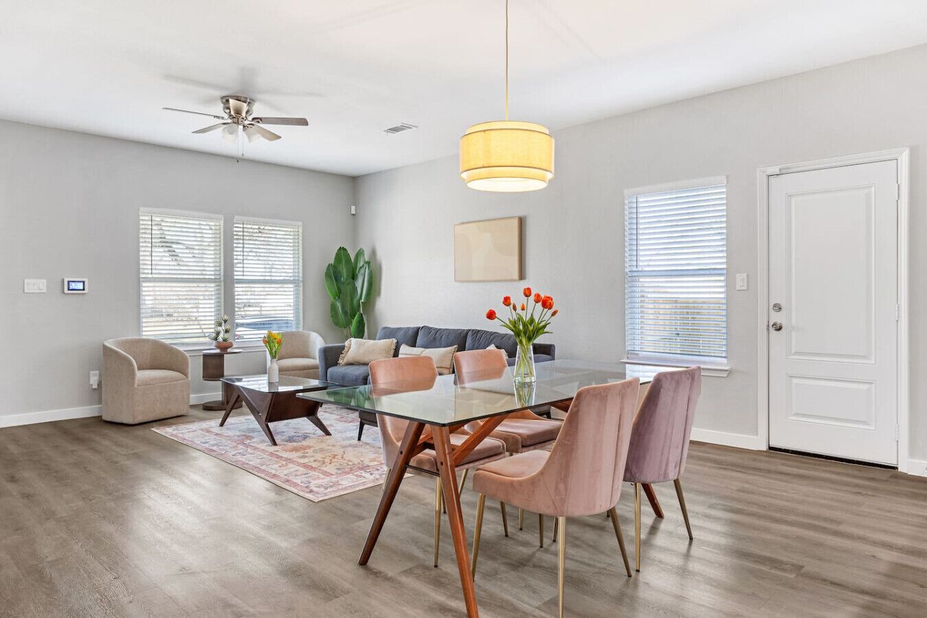 A modern living room with a glass dining table, comfortable seating, a ceiling fan, and large windows with blinds.