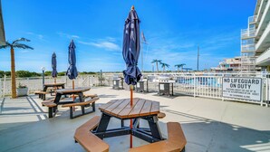 A deck with picnic tables and umbrellas, a lifeguard duty sign, and a view of the water and buildings. - Beachfront Condo in Wildwood Crest w/ Pool Access! (Wildwood Crest)