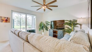 A modern living room with a large beige sectional sofa, a wooden ceiling fan, a large window with a view of trees, and a potted plant. - Ibis Landing Golf Club On-Site: Condo w/ Porch! (Lehigh Acres)