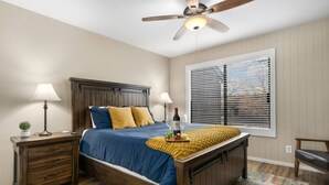 A bedroom with a bed, wooden headboard, bedside tables, lamps, a ceiling fan, a window with blinds, and a chair. - County Club Townhome *Packman Game, Xbox, Golf course Views, Near Forest Edge (Flagstaff)