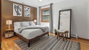 Ein Schlafzimmer mit Bett, Nachttischlampen, Teppich, Fenster mit Vorhängen, Deckenventilator und einem großen Spiegel.