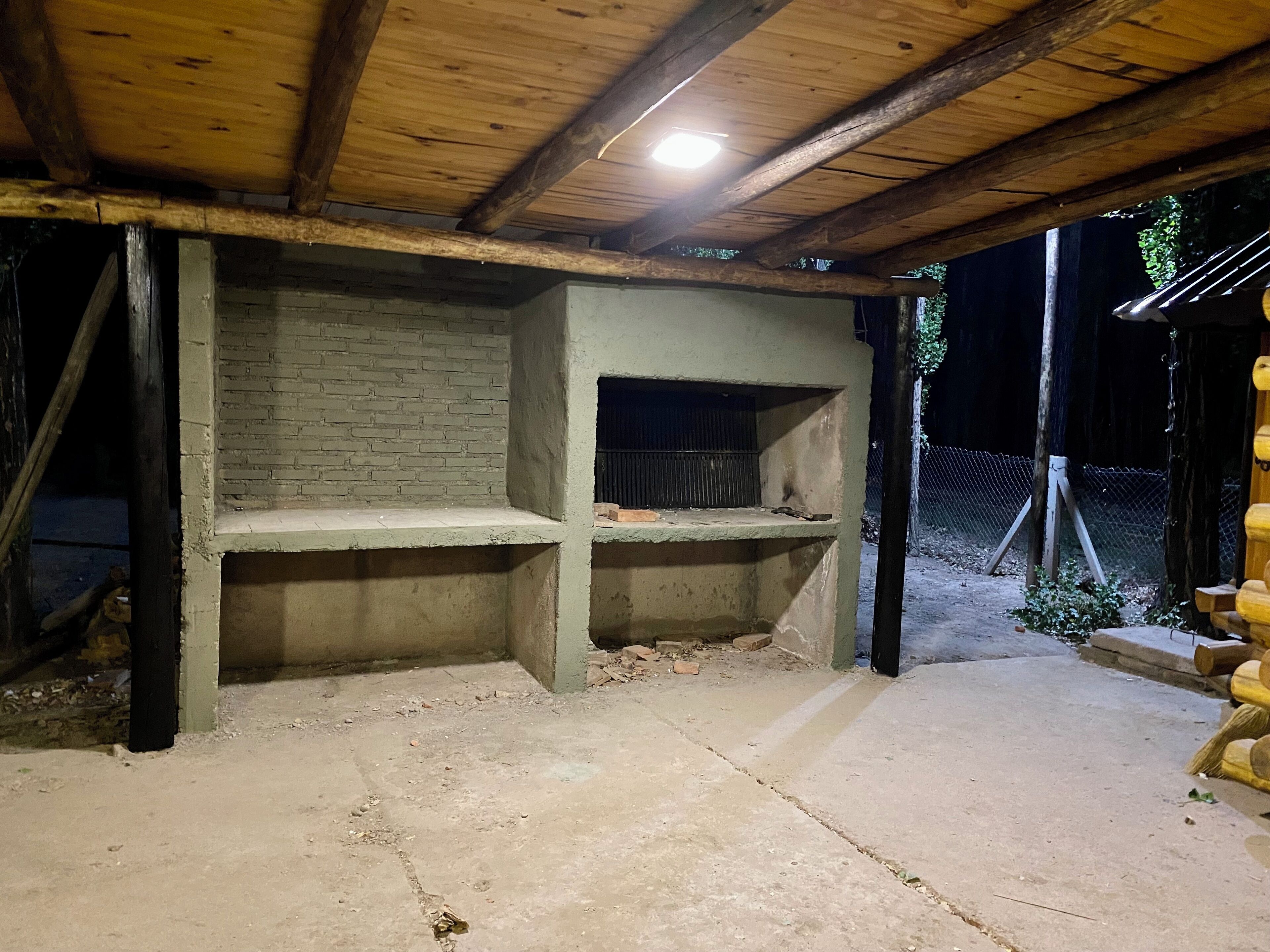Una estructura rústica al aire libre con pared de ladrillo y piso de concreto, que incluye un estante metálico y techo con vigas de madera.