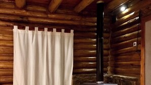 A log cabin interior with a wood-burning stove, wooden walls, a white curtain, and a tiled floor.
