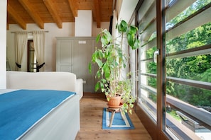A room with a bed, a potted plant, a window with a view of greenery, and a wooden ceiling.