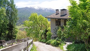 Ein rustikales Haus mit einem Feldweg, der davor hindurchführt, umgeben von üppiger Vegetation und Bergen im Hintergrund.
