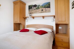 A bedroom with a bed, two red pillows, a wooden headboard, a nightstand, a wall-mounted shelf, and a painting of a rural scene.
