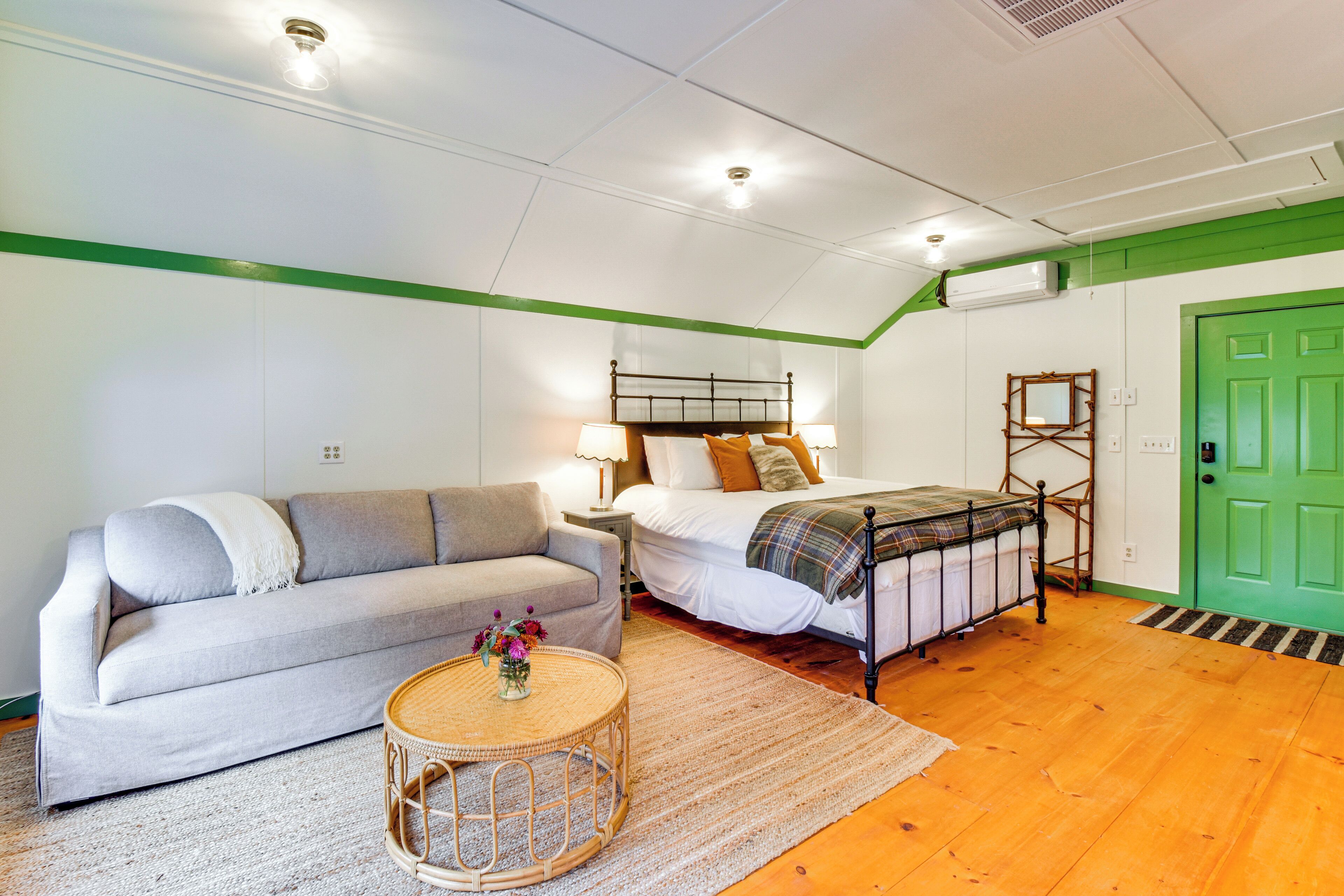 A bedroom with a bed, a sofa, a bedside table with lamps, a green door, and a wooden floor.