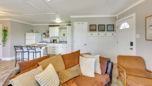Een woonkamer met een bruine bank, witte kussens en een rieten stoel. Achtergrond toont een keuken met witte kasten en een eethoek.