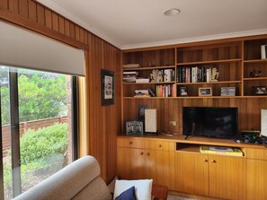 Una sala de estar con un televisor grande, estanterías de madera y una ventana con vistas a la vegetación.