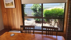 Une pièce avec une table en bois et deux chaises, une fenêtre donnant sur un jardin et un bâtiment à l’extérieur, et un tableau encadré accroché au mur.