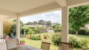 Une terrasse couverte avec une table et des chaises, une vue sur un jardin bien entretenu et, au loin, un quartier résidentiel.