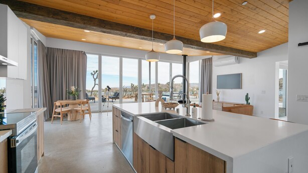 A modern kitchen with a large island, wooden ceiling, and large windows offering a view of the desert. - Alto Yucca- Joshua Tree Modern Glass house with private pool (Yucca Valley)