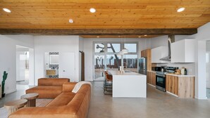 A spacious living area with a wooden ceiling, a large window, and a kitchen with modern appliances. - Alto Yucca- Joshua Tree Modern Glass house with private pool (Yucca Valley)