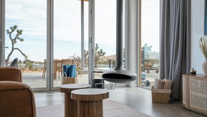 A modern living room with a large glass door leading to a patio area. There are wooden tables and chairs, a hanging light fixture, and a wicker basket with towels. - Alto Yucca- Joshua Tree Modern Glass house with private pool (Yucca Valley)
