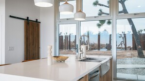 A modern kitchen with a large island, a wooden slat wall, and a view of a desert landscape through large windows. - Alto Yucca- Joshua Tree Modern Glass house with private pool (Yucca Valley)