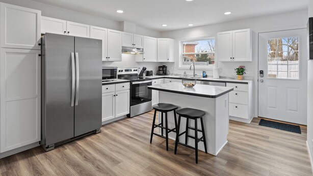 A modern kitchen with white cabinets, stainless steel appliances, and a central island with stools. - Modern Waters- Bright and Central- Walk to Downtown (Lake Geneva)