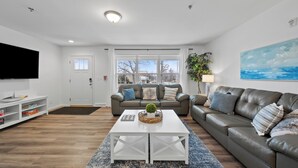 A modern living room with a white coffee table, a grey sofa, a television, and a large window. - Modern Waters- Bright and Central- Walk to Downtown (Lake Geneva)
