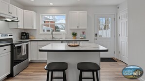 A modern kitchen with white cabinets, a central island with two stools, a stainless steel oven, and a window providing natural light.