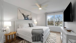 A modern bedroom with a large bed, a ceiling fan, a TV, and a window with a view of the outside. - The Blackstone| Arcade and Game Lounge|Near Downtown|New Construction|Groups Welcome! (Lake Geneva)