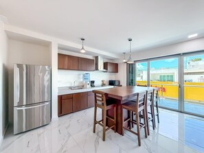 A modern kitchen with a stainless steel refrigerator, wooden cabinets, a marble countertop, and a dining area with a table and chairs. - 2BR Condo w/Rooftop Pool (Isla Mujeres)