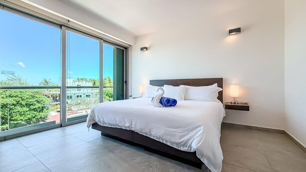 A modern hotel room with a large bed, a view through a sliding glass door, and a small bedside table with a lamp. - 1BR w/Rooftop Pool Downtown (Isla Mujeres)
