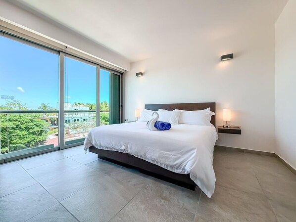 Una habitación de hotel moderna con una cama grande, vista a través de un ventanal corredero y una mesita de noche pequeña con lámpara.