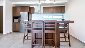 A modern kitchen with wooden cabinets, a stainless steel refrigerator, and a dining area with wooden chairs. - 1BR w/Rooftop Pool Downtown (Isla Mujeres)
