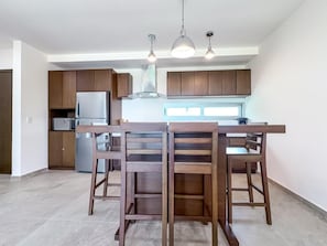 A modern kitchen with wooden cabinets, a stainless steel refrigerator, and a dining area with wooden chairs. - 1BR w/Rooftop Pool Downtown (Isla Mujeres)