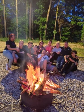 Eine Gruppe Menschen sitzt bei einem Lagerfeuer in einer natürlichen Umgebung, im Hintergrund sind Bäume zu sehen.