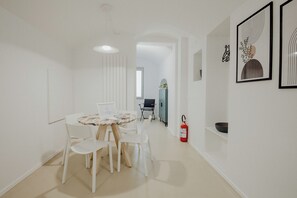 Un espace repas moderne et minimaliste, avec une table ronde et des chaises blanches, un extincteur fixé au mur et des œuvres d’art encadrées qui décorent le mur.