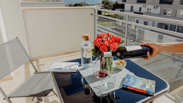 A balcony with a table set for two, featuring a bottle of wine, a glass, a vase with red flowers, a book, and a water bottle. - Rainbow Relax Home by MONHOLIDAY (Monopoli)