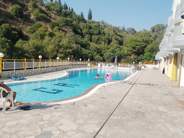 Una piscina con terraza de piedra, rodeada por una cerca amarilla y blanca, y un terraplén al fondo.