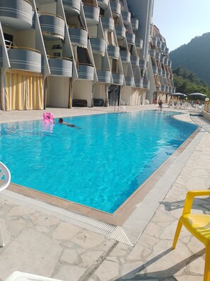 Una piscina con borde de baldosas, rodeada por un edificio con balcones y una montaña al fondo.
