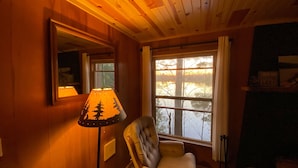Ein gemütliches Zimmer mit Holzwänden, einem großen Fenster mit Vorhängen, einem Sessel und einer Lampe mit Waldmotiv.