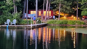 Un chalet au bord du lac, doté d’un quai, entouré de grands arbres et bordé par une étendue d’eau calme où se reflètent les lumières.