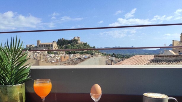 Ein Frühstücksbrett mit Croissant, Brot, Ei und Saft auf einem Balkon mit Blick auf eine Stadtlandschaft und historische Gebäude.