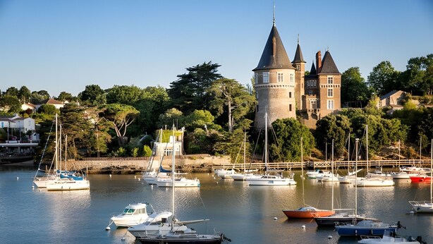 Uma vila costeira com uma torre e castelo imponentes, rodeada por uma marina com diversos barcos.