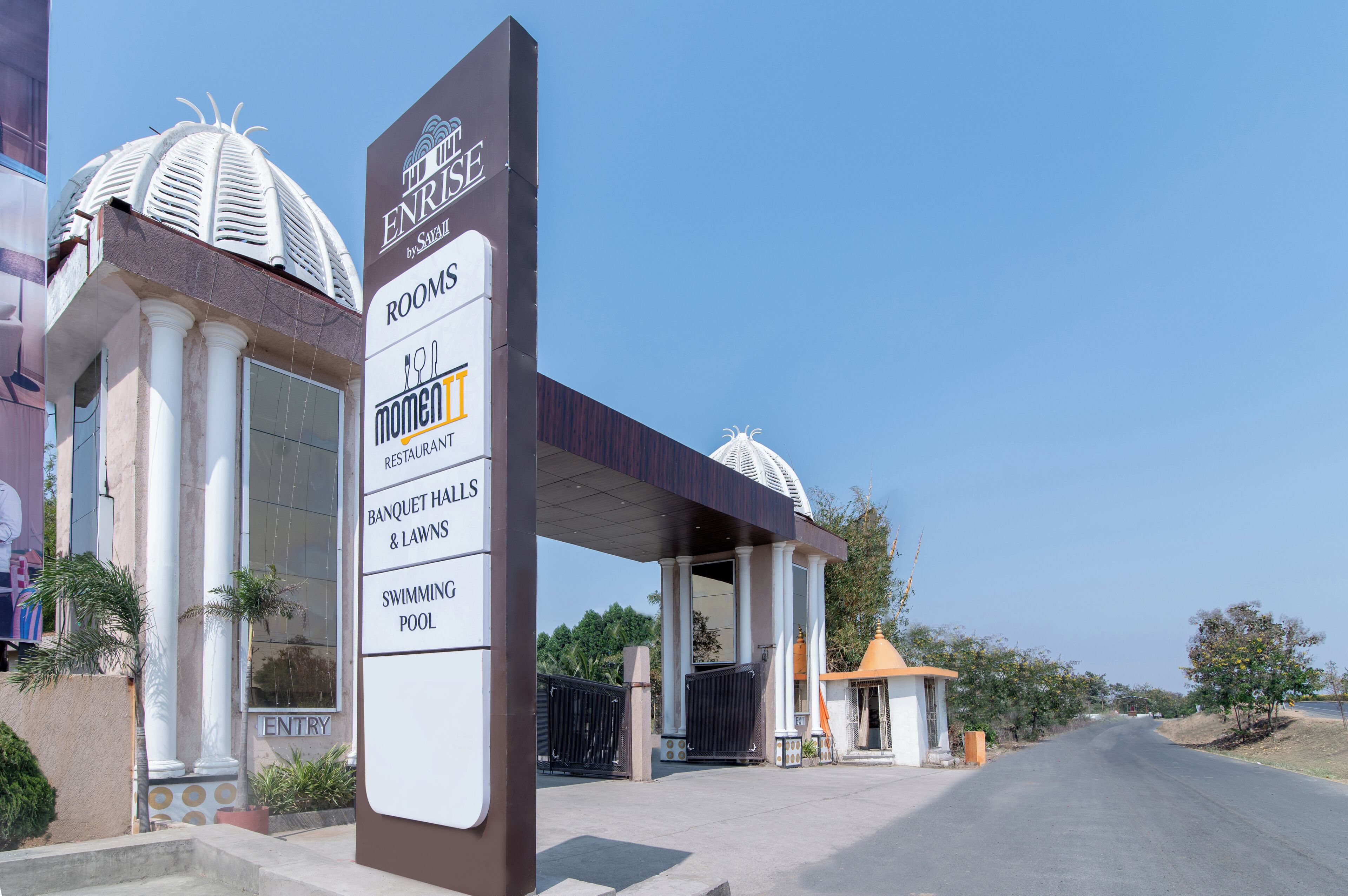 A hotel entrance with a sign listing amenities including rooms, a restaurant, banquet halls, lawns, and a swimming pool.