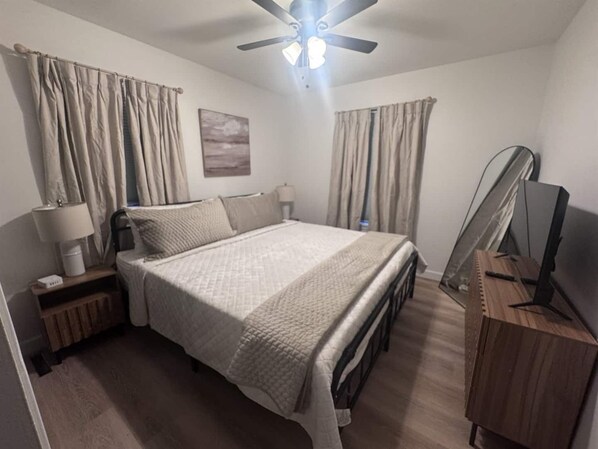 Une chambre à coucher comprenant un lit, un bureau, une chaise, un ventilateur de plafond et des rideaux.