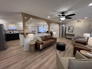 A spacious living area with a sofa, a coffee table, and a dining area with a kitchen in the background. - Organic Oasis - Spa Arcade (Maize)