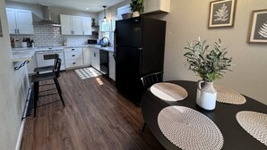 A compact kitchen with white cabinets, a black refrigerator, and a dining area with a black table and woven placemats. - Radian Ranch - 2 Kings, Near Derby - Fenced Yard - Pet Friendly. (Wichita)