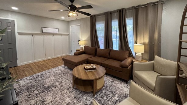 A modern living room with a brown sectional sofa, a round coffee table, and a beige armchair. There is a ceiling fan, a window with curtains, and a door with a coat of arms. - Radian Ranch - 2 Kings, Near Derby - Fenced Yard - Pet Friendly. (Wichita)