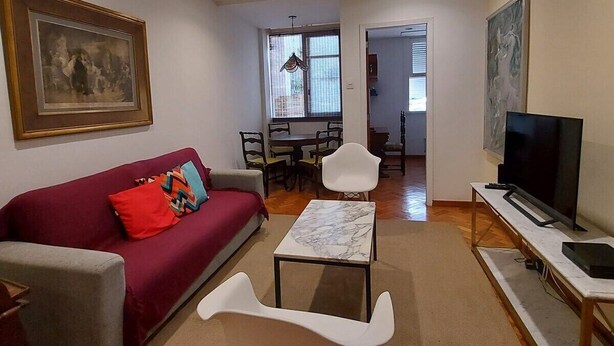 Una sala de estar con un sofá rojo, un sillón blanco, una mesa de centro de mármol, un televisor sobre un mueble bajo y una zona de comedor al fondo.