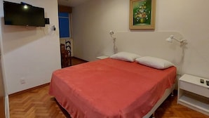 Un dormitorio con una cama, televisor montado en la pared, un cuadro sobre la cama y una mesita de noche blanca.