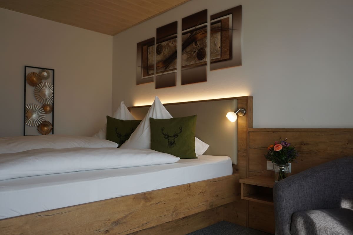 Ein Schlafzimmer mit einem hölzernen Kopfstück, einem Bett mit weißer Bettwäsche, dekorativen Kissen, einer Wandleuchte und einem kleinen Regal mit einem Blumenstrauß.